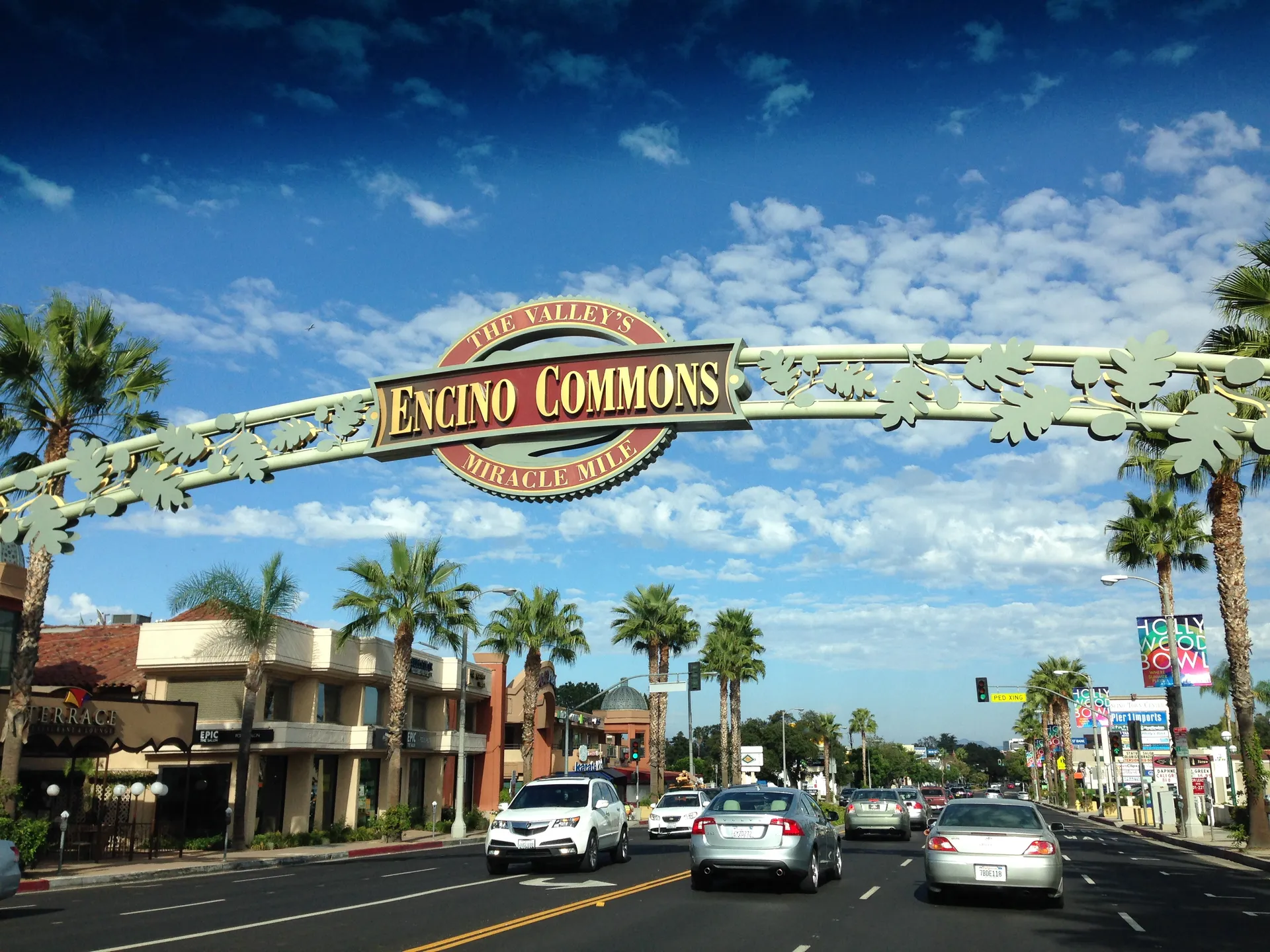 Cars pass beneath the Encino Commons arch over Ventura Boulevard, with shops and palm trees on either side.