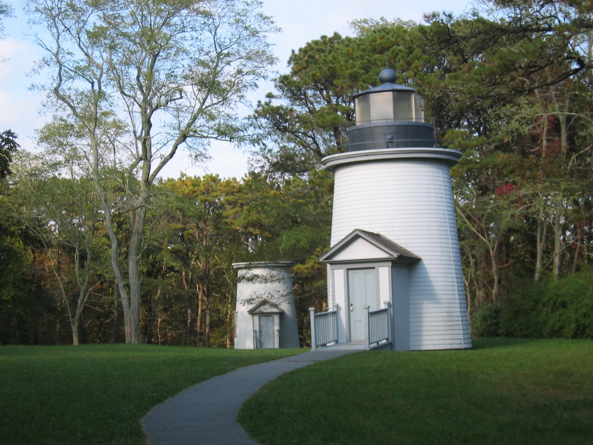Two of the Three Sisters of Nauset lighthouses in North Eastham, small white towers, stand among pines along a curving footpath.