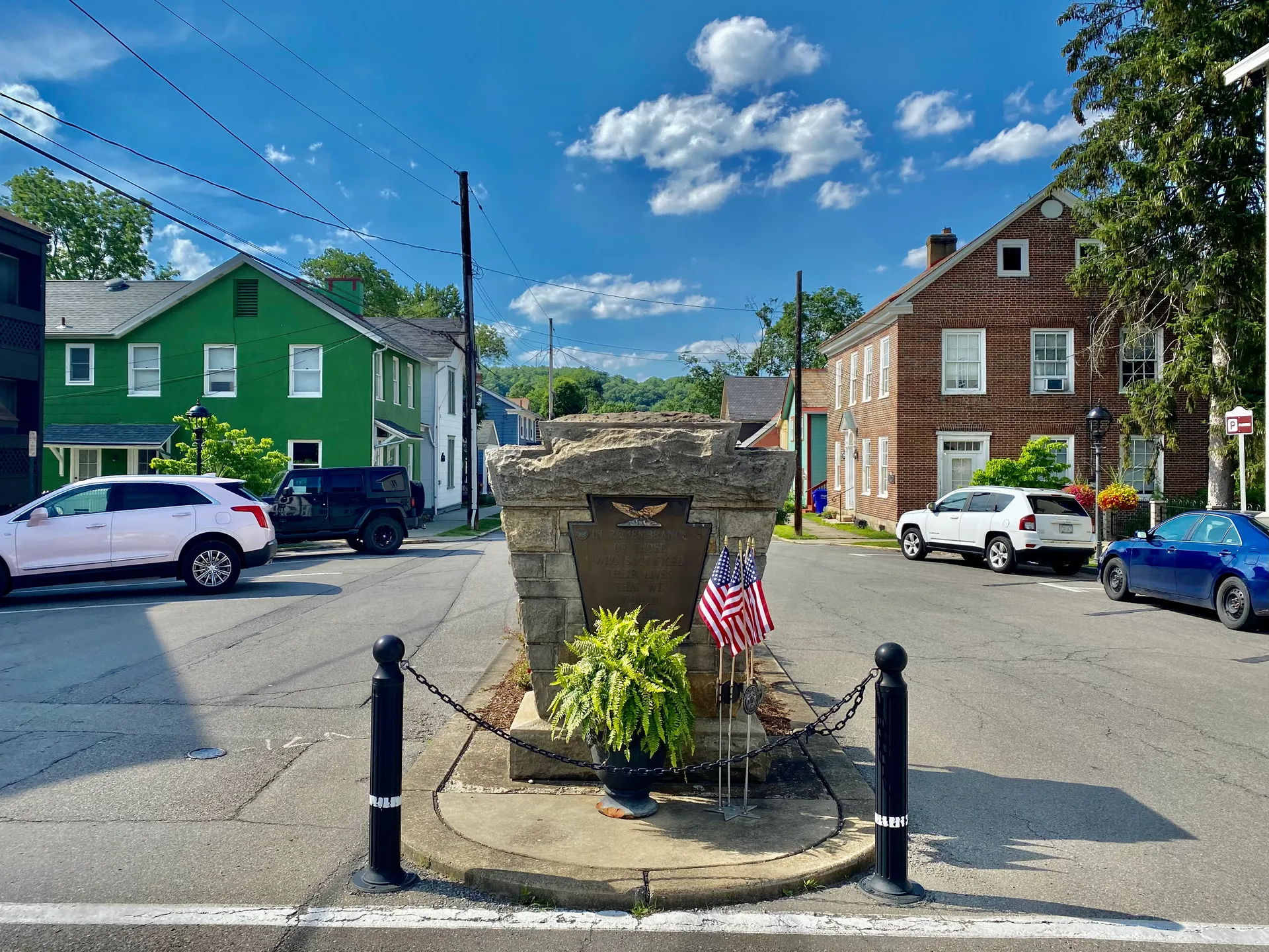 A keystone-shaped veterans monument with flags and flowers anchors a small plaza on Main Street in Harmony, near a bright green building.
