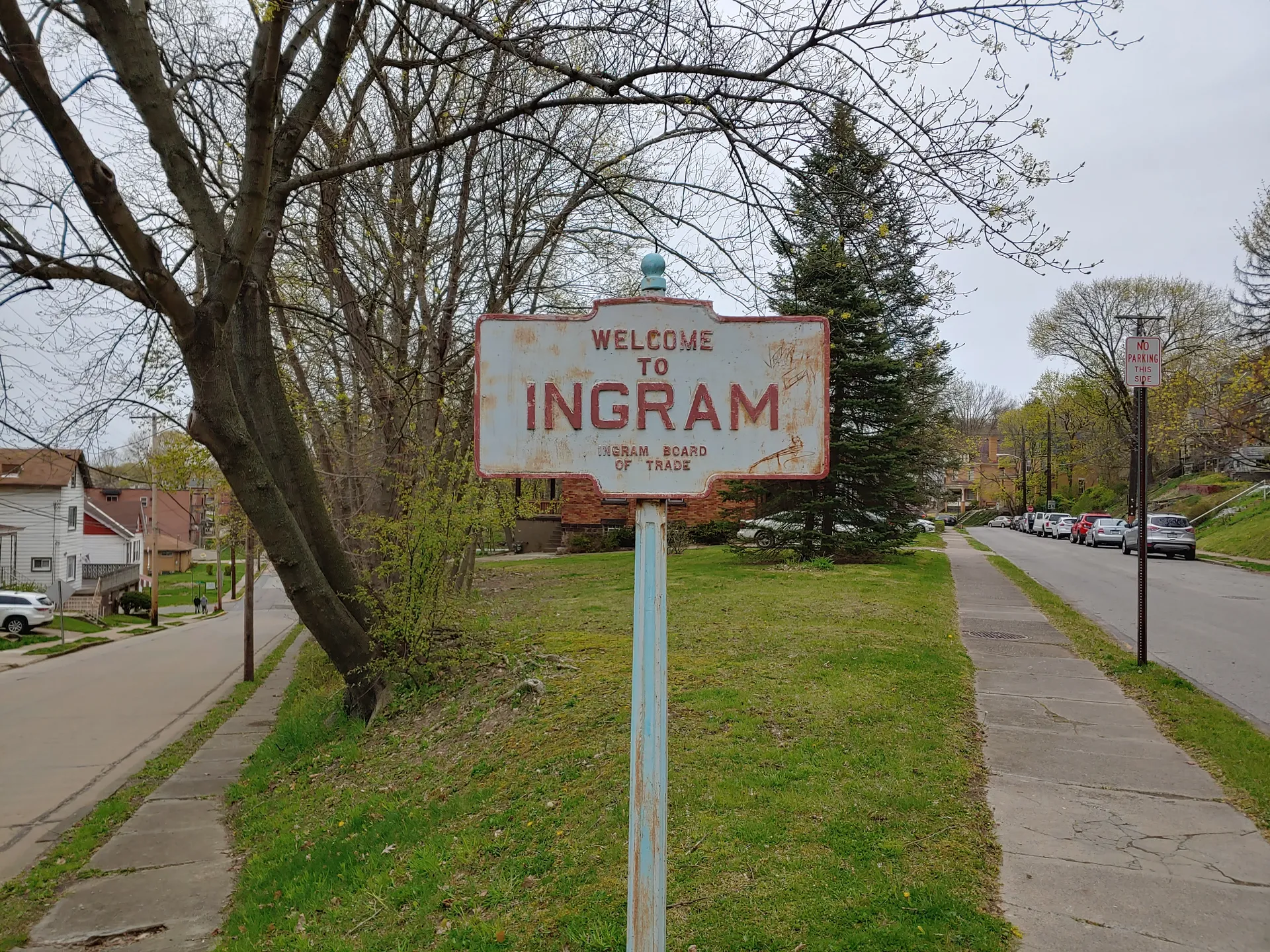 A white-and-red welcome sign for Ingram stands on a grassy strip beside a sidewalk lined with homes and autumn trees.