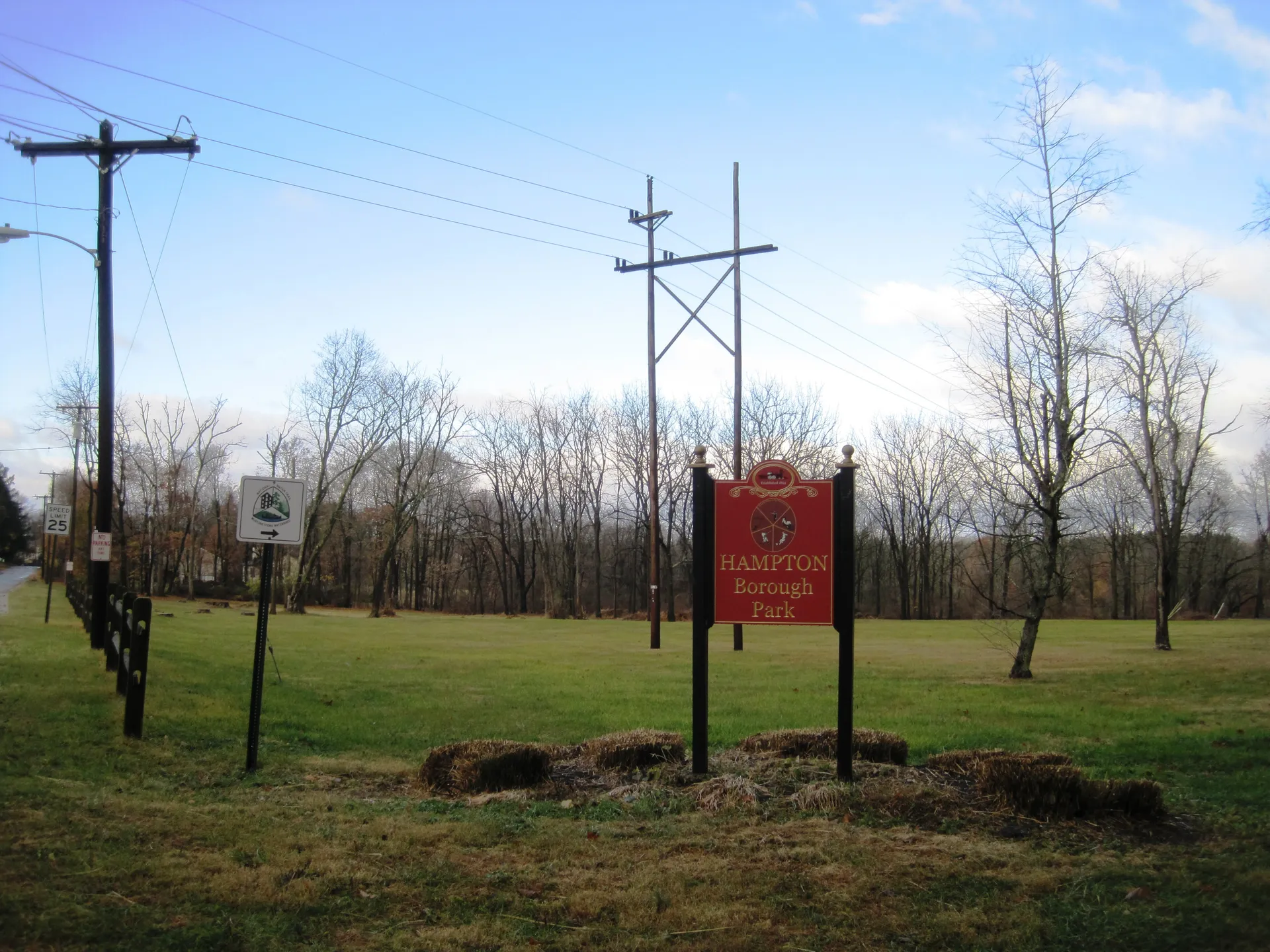 The entrance to Hampton borough park is marked by a red sign on posts, with an open lawn and woods behind it.
