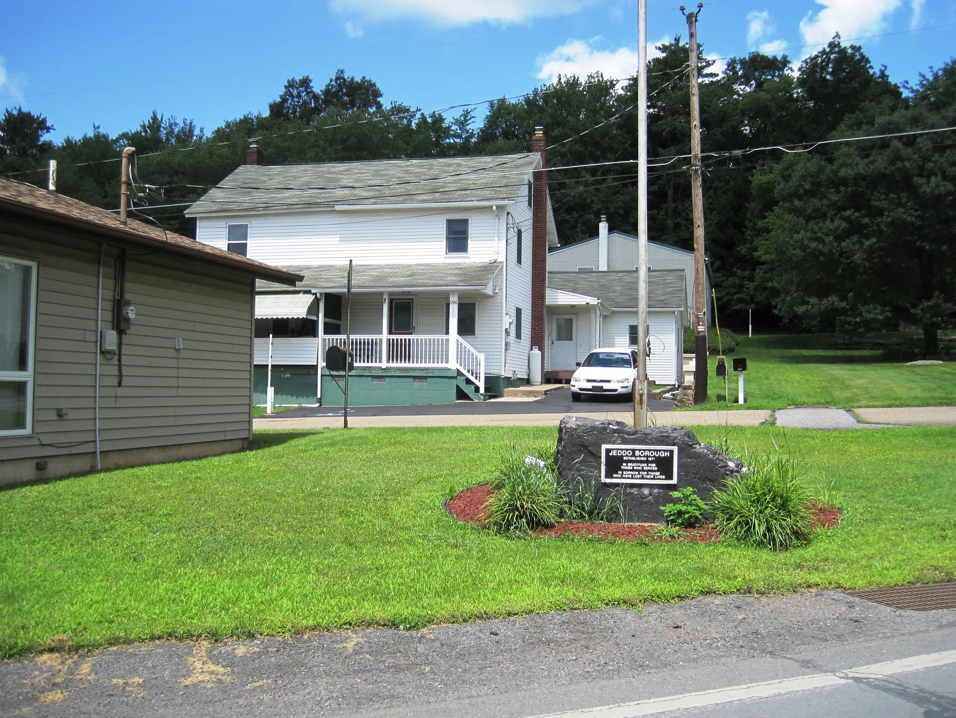 A veterans memorial rock and plaque sit in a flower bed on a small green in Jeddo, with houses behind it.