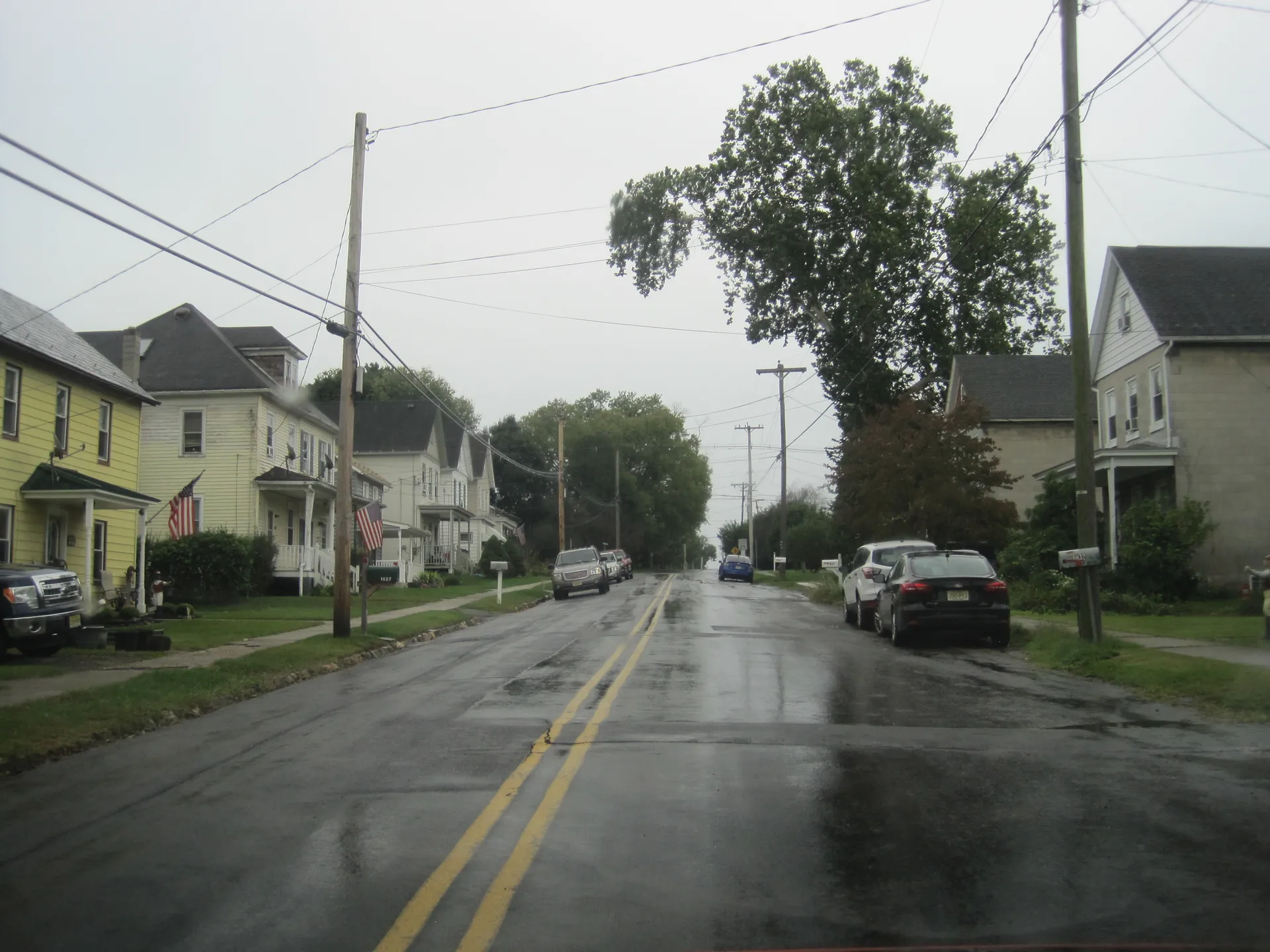 A rainy residential street in Brainards curves past older frame houses and parked cars beneath tall shade trees.