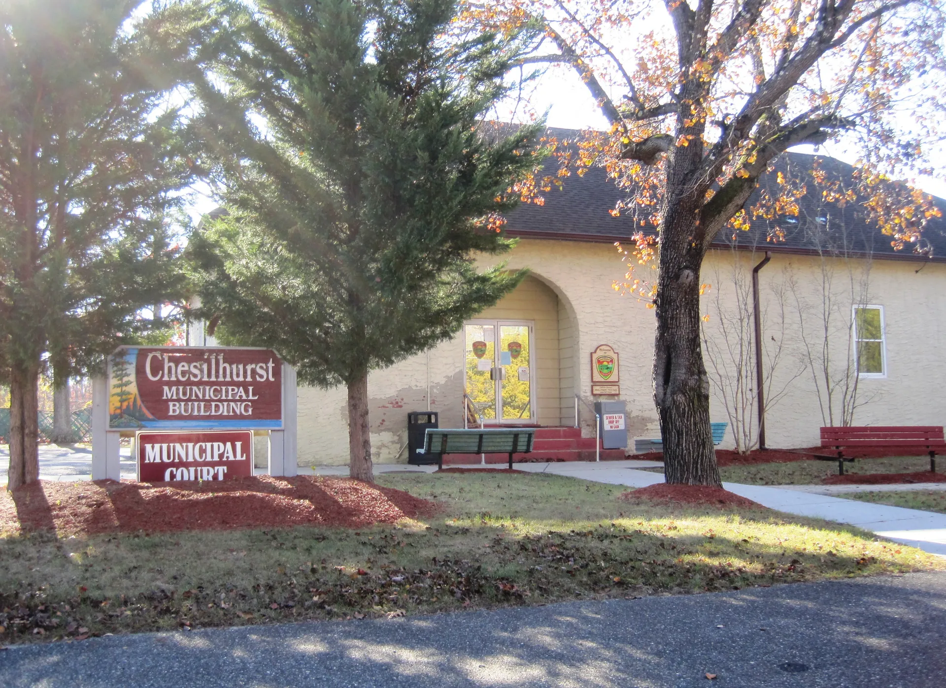 Chesilhurst's municipal building is a cream stucco structure with a red sign out front, partly shaded by a large evergreen.