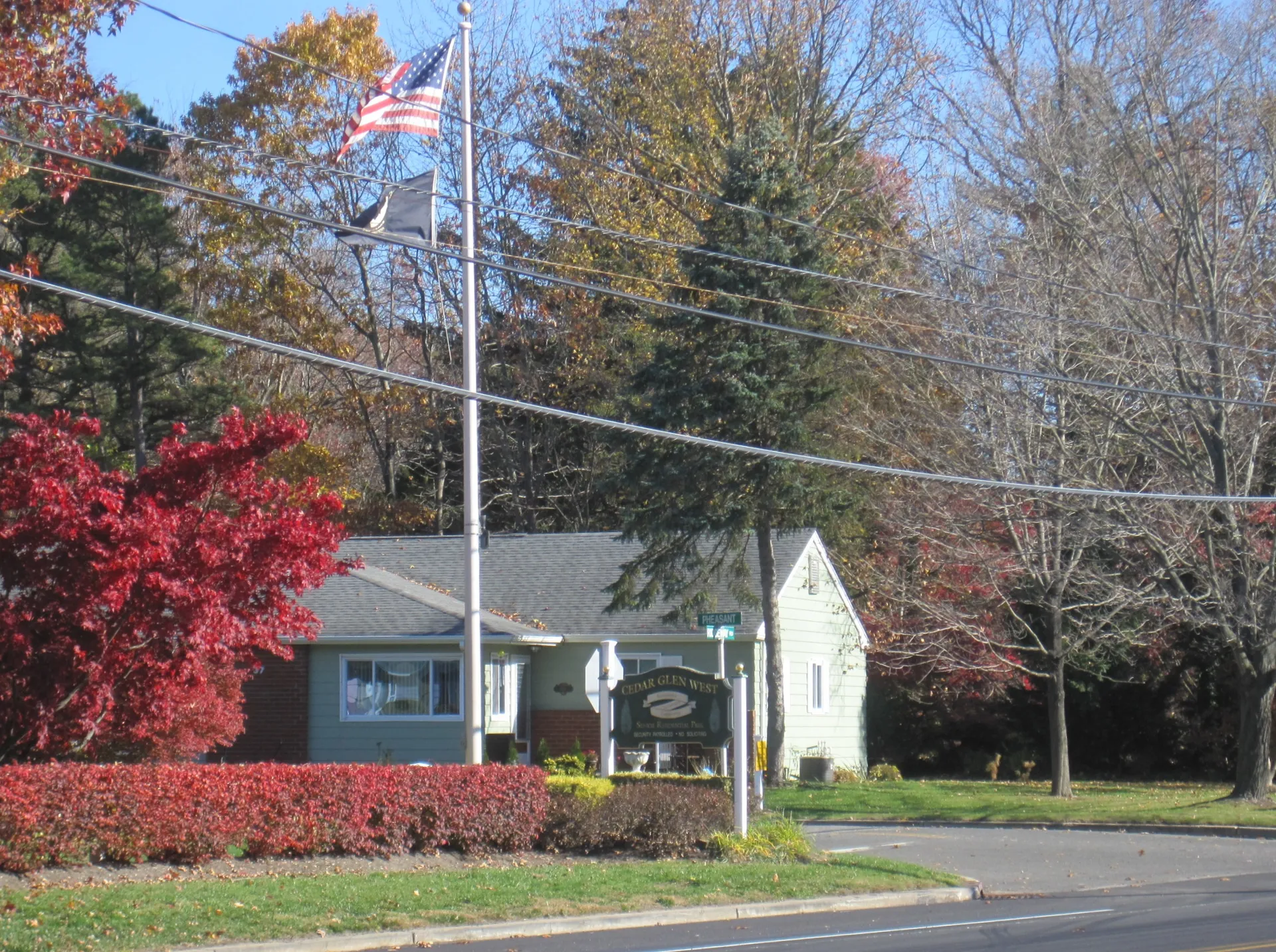 At the entrance to Cedar Glen West, a flag flies over a sign and a small gray clubhouse framed by red shrubs and autumn trees.