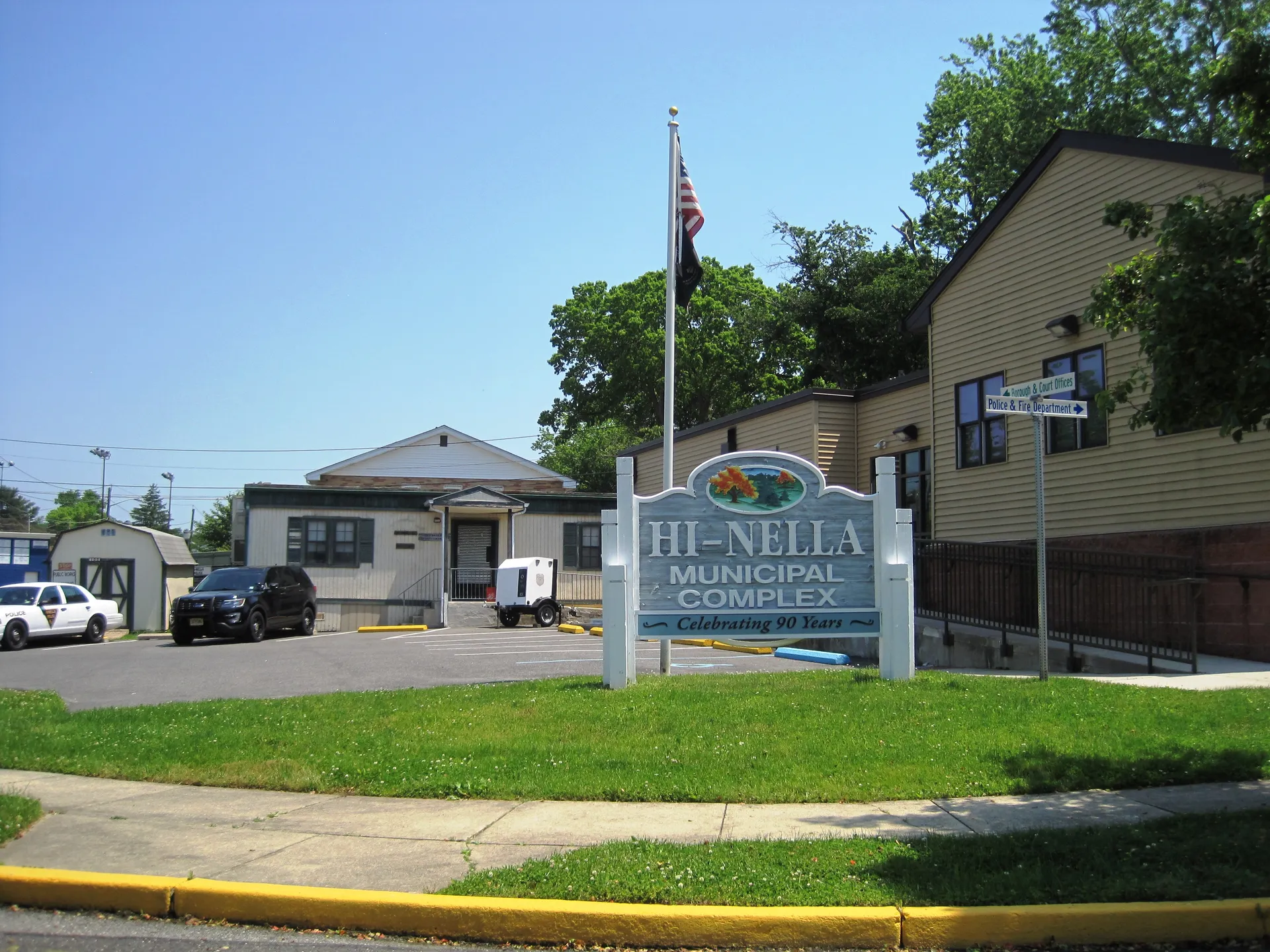 The Hi-Nella municipal complex is a small building with a flag and a sign set in a landscaped bed on the lawn.