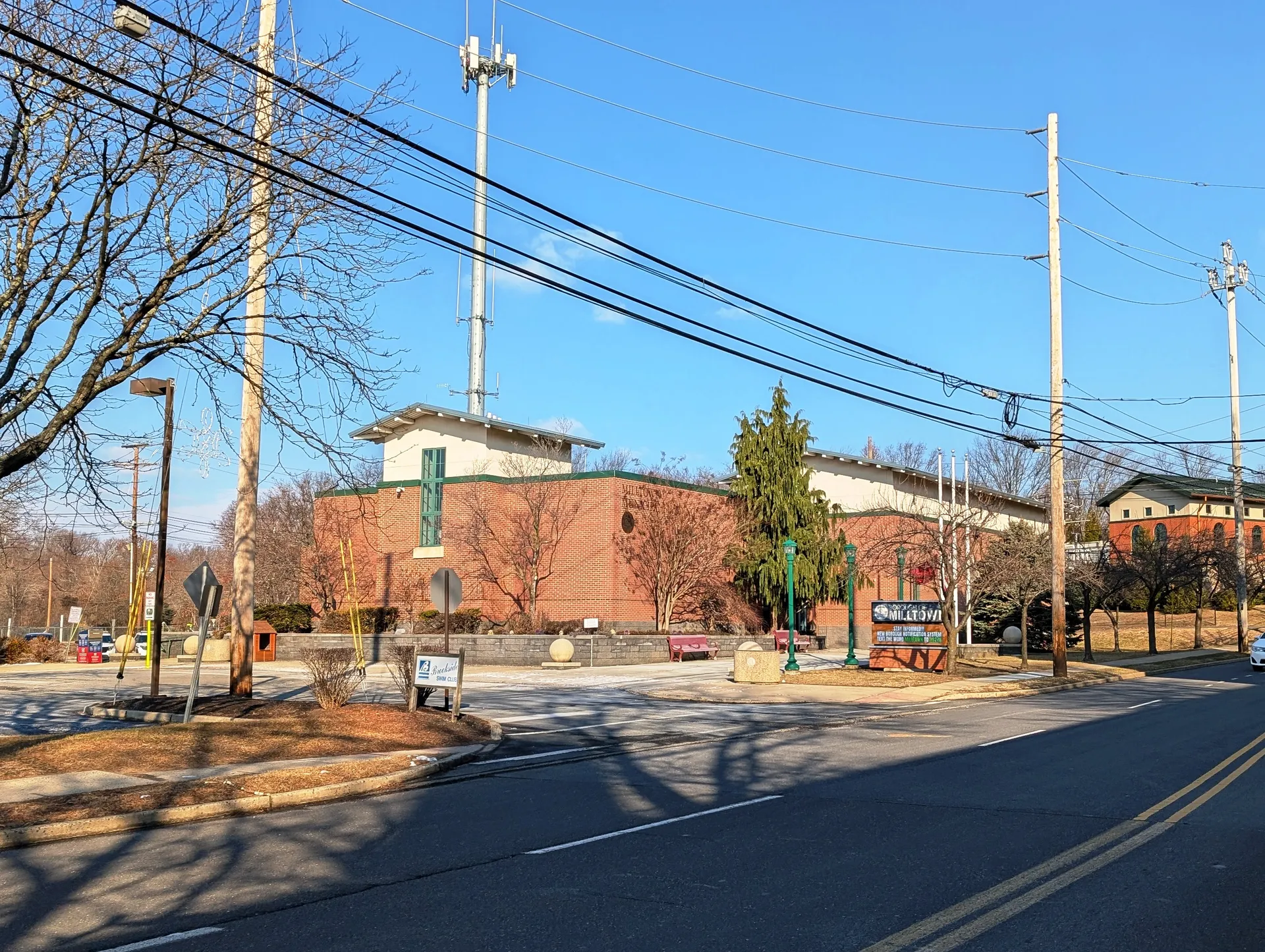 The Milltown municipal building is a red brick building with a flag, seen past a mulched corner garden.