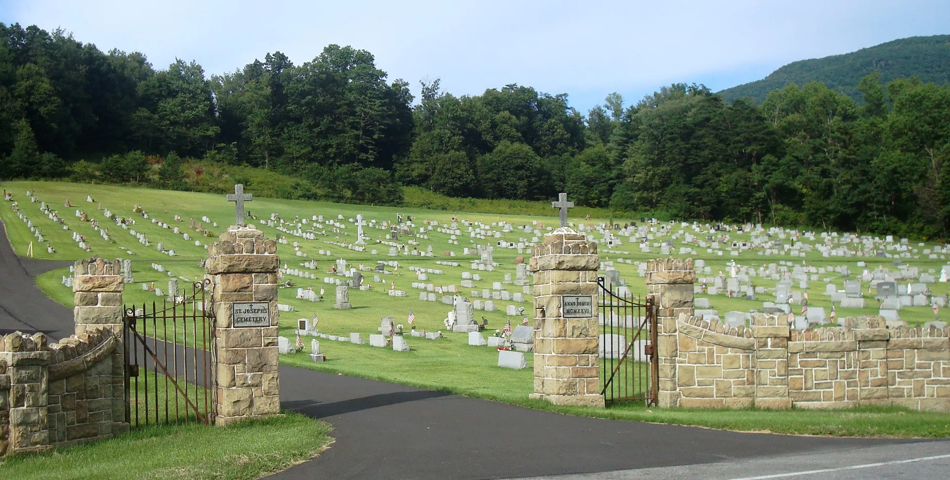 Stone pillars and an iron gate open onto St. Joseph's Cemetery, its headstones climbing a green slope beside the Bucktail Trail in Hyner.