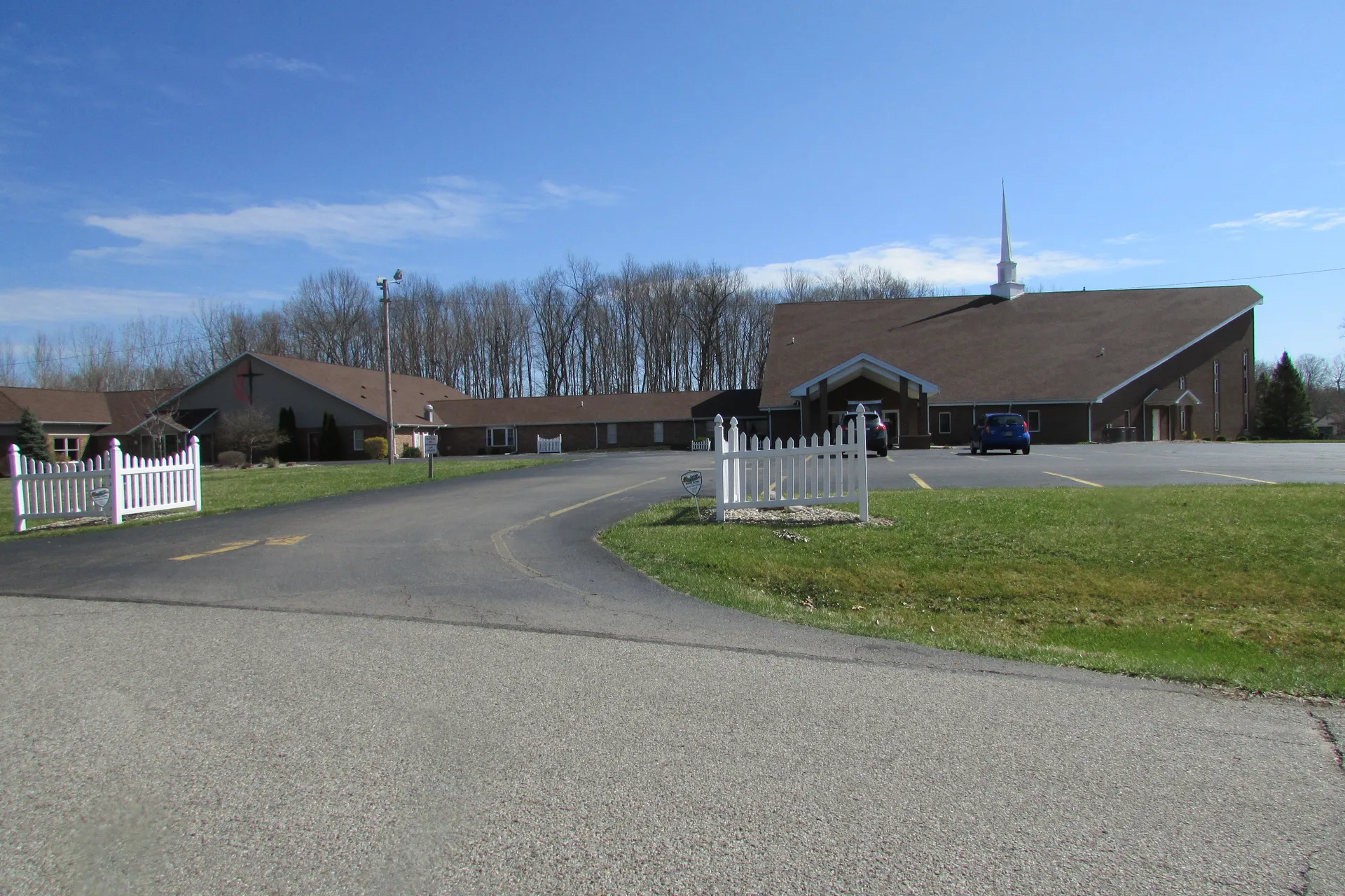 A paved drive curves toward a long, low brick church in North Webster, past a white picket fence section and mowed grass.
