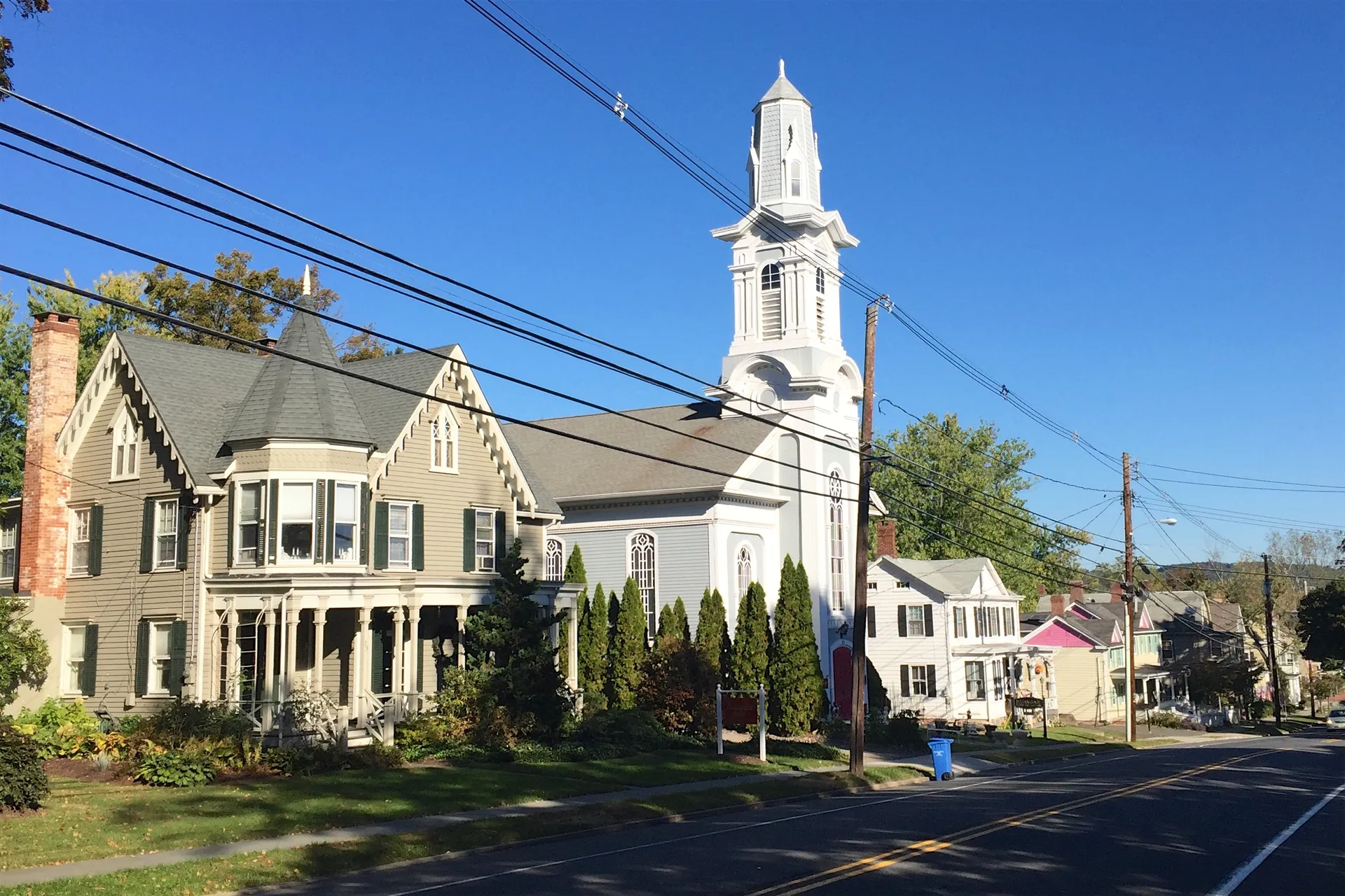 Looking north along Main Street in Oldwick, a white church steeple rises behind clapboard houses and trimmed shrubs.
