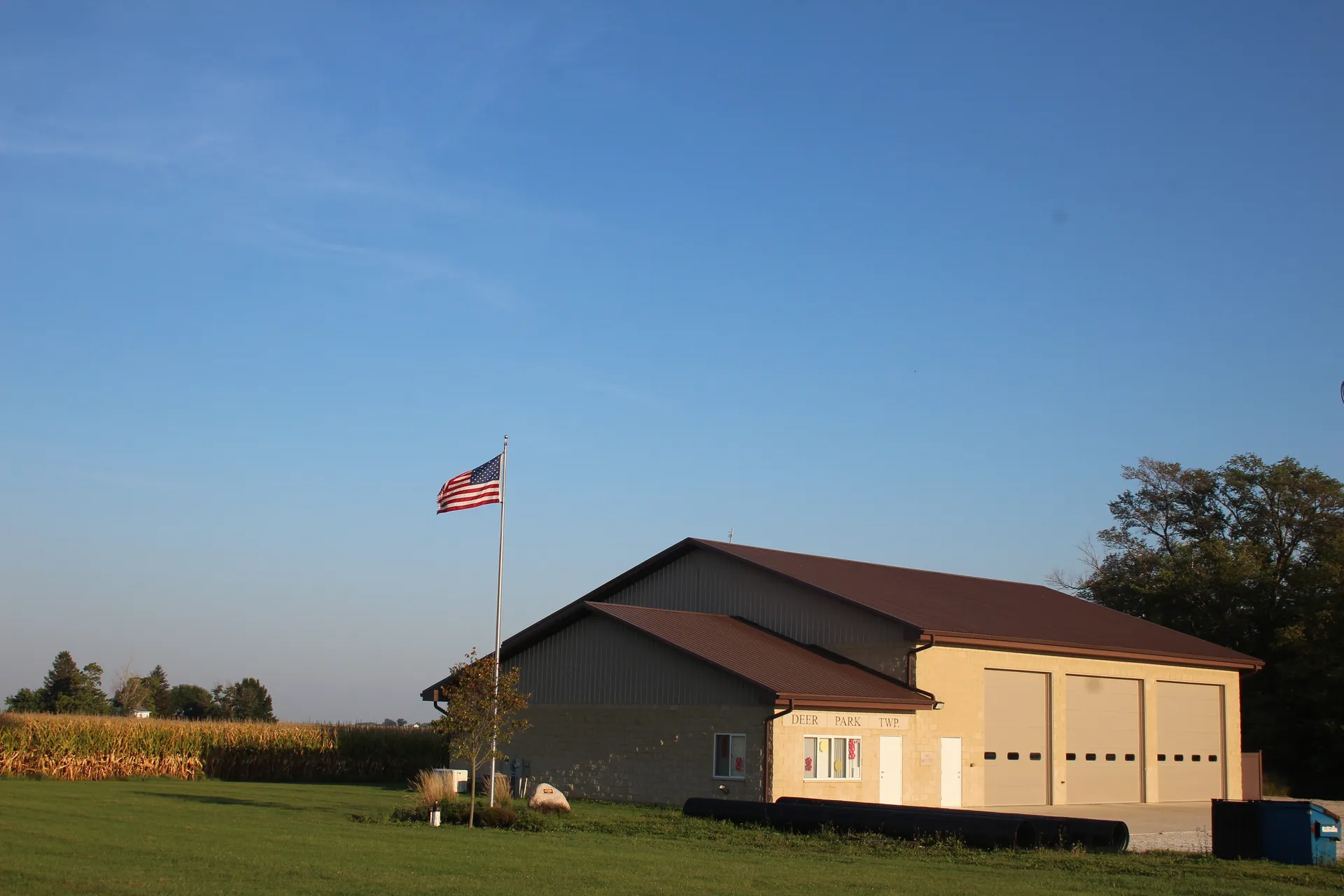 An American flag flies beside the Deer Park Township building, a tan steel structure with a dark red roof on a grassy rise.