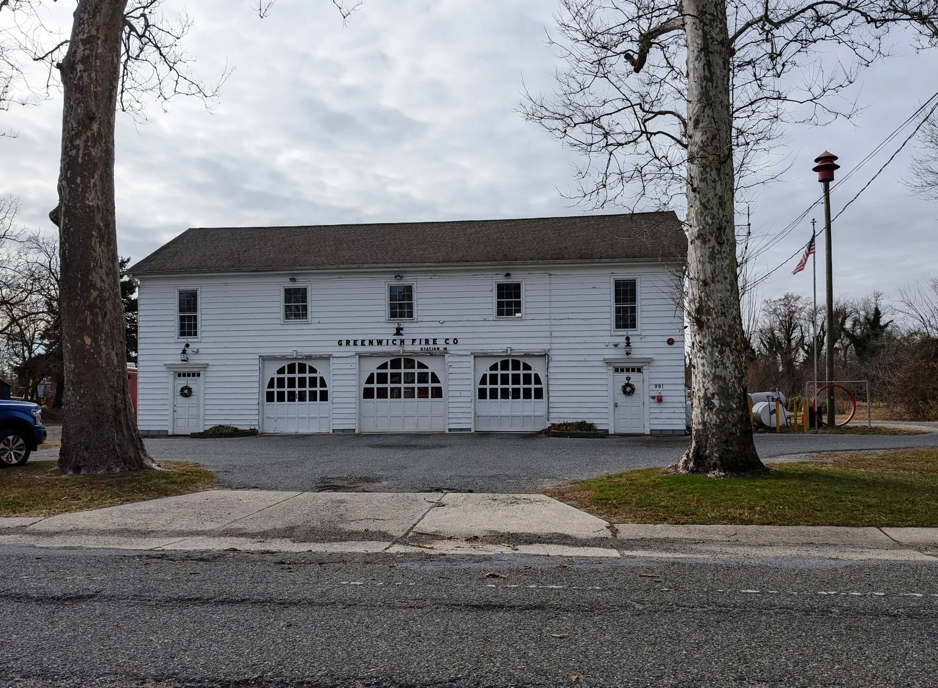 The Greenwich Fire Company firehouse is a white building with arched bay doors and a gabled roof under tall trees.