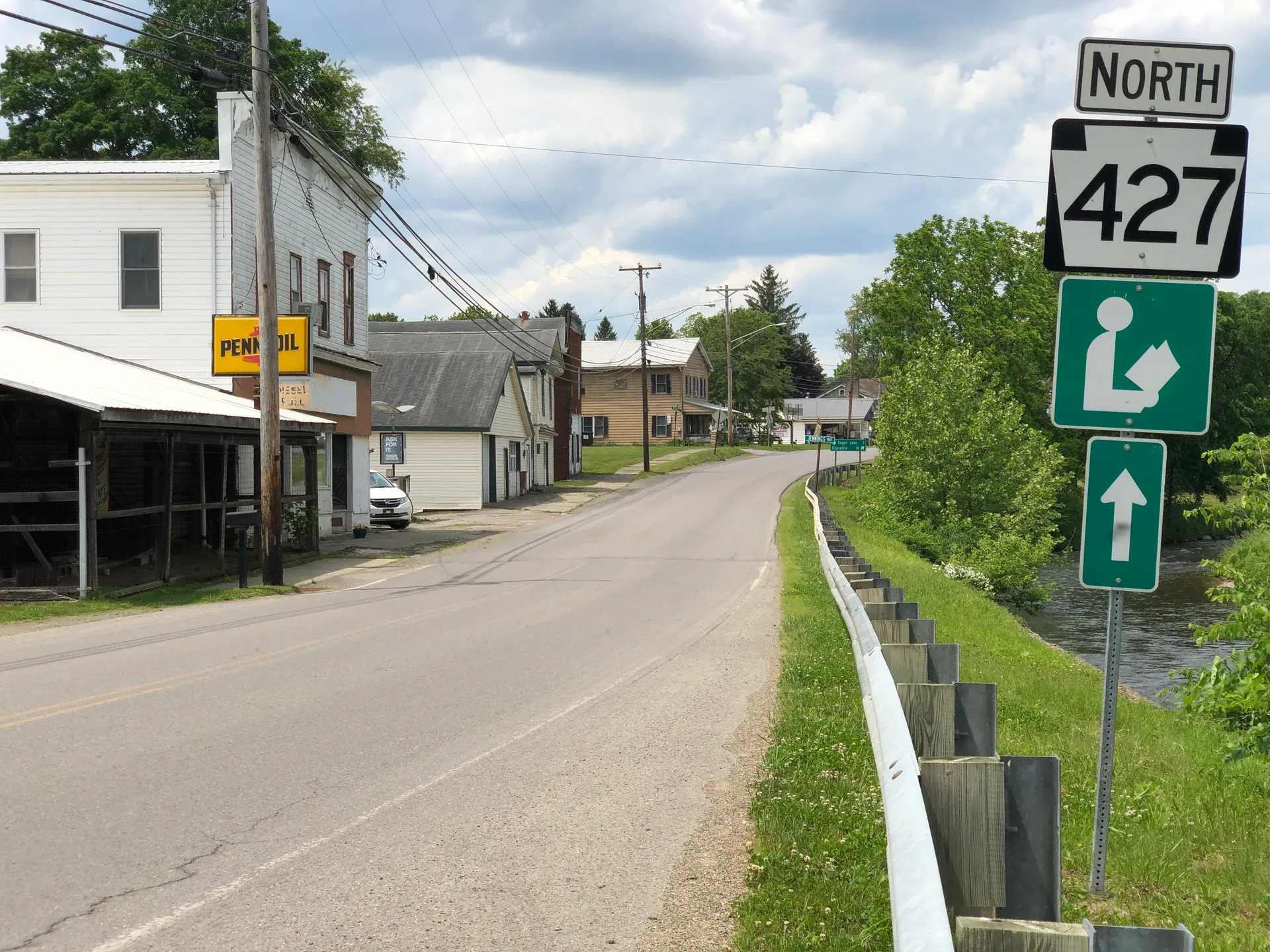 Main Street (Route 427) in Cooperstown passes Factory Street, flanked by older frame houses and a guardrail along the creek.