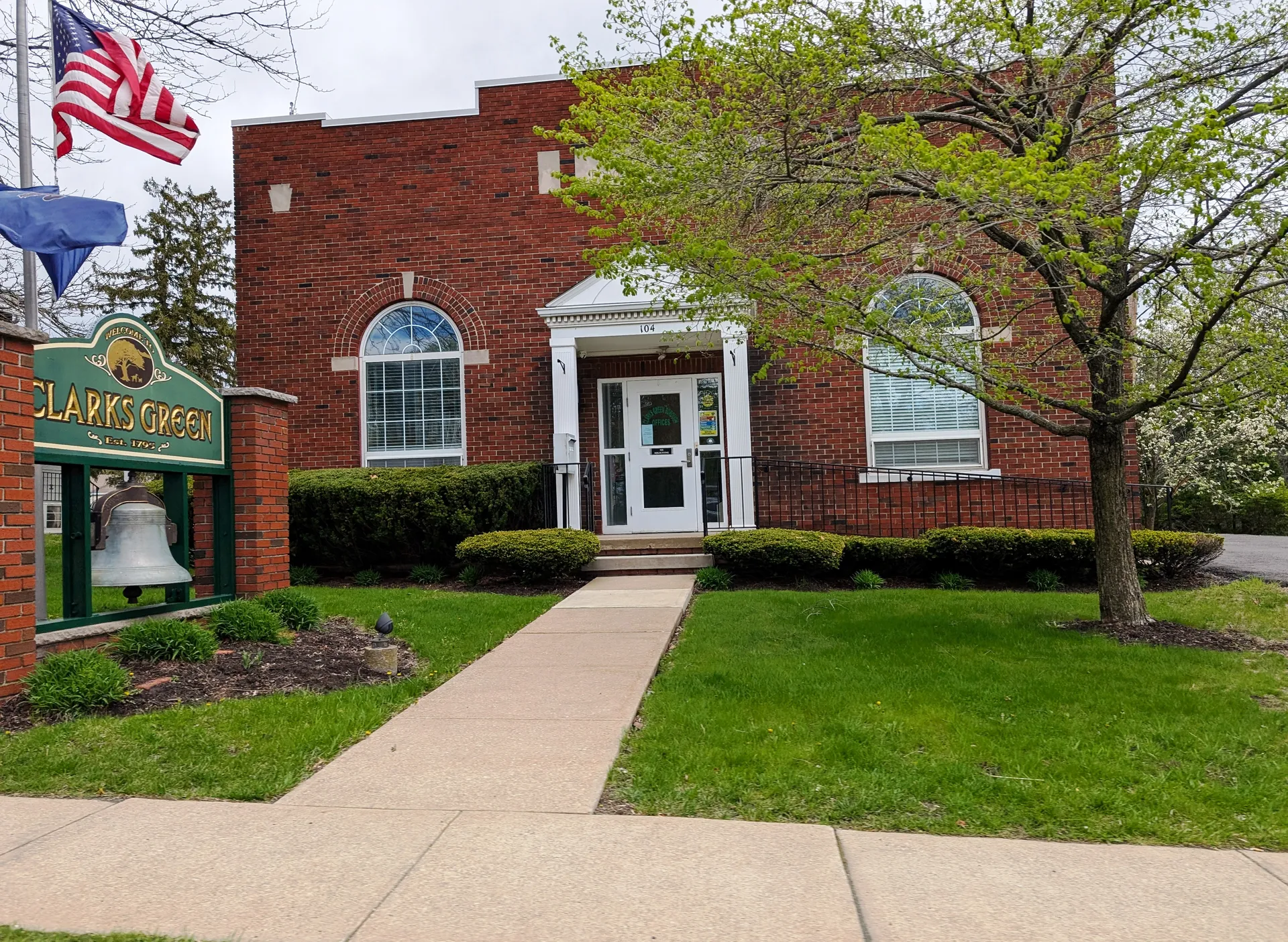 A green sign with a bell marks the Clarks Green Borough Offices, a red-brick building with a white portico and arched window.