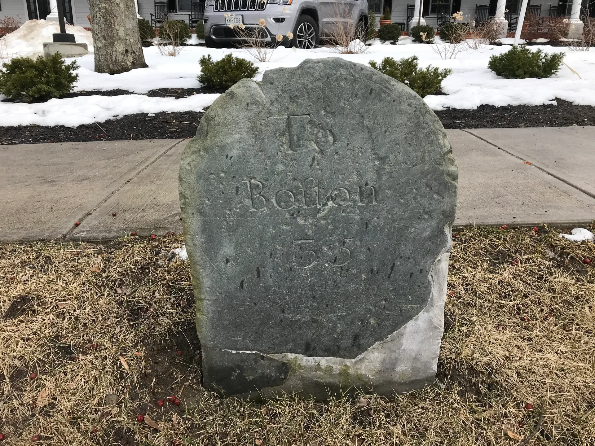 A weathered granite milestone in Groton is carved with the distance to Boston, set in a mulched bed beside the road.