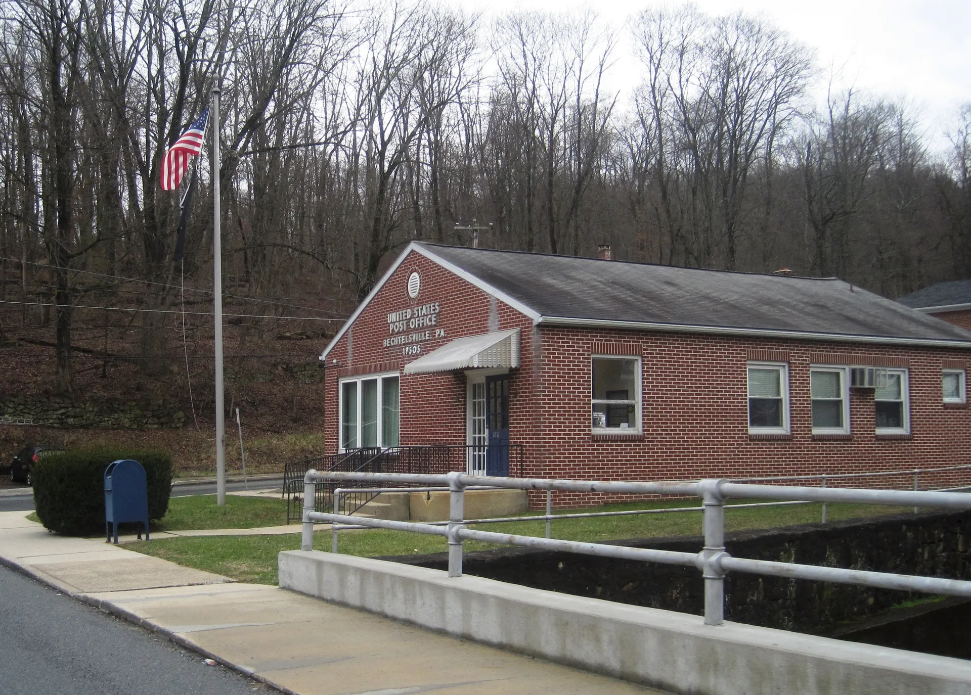 Bechtelsville's post office is a compact brick building with a white railing, seen from North Main Street against a wooded backdrop.