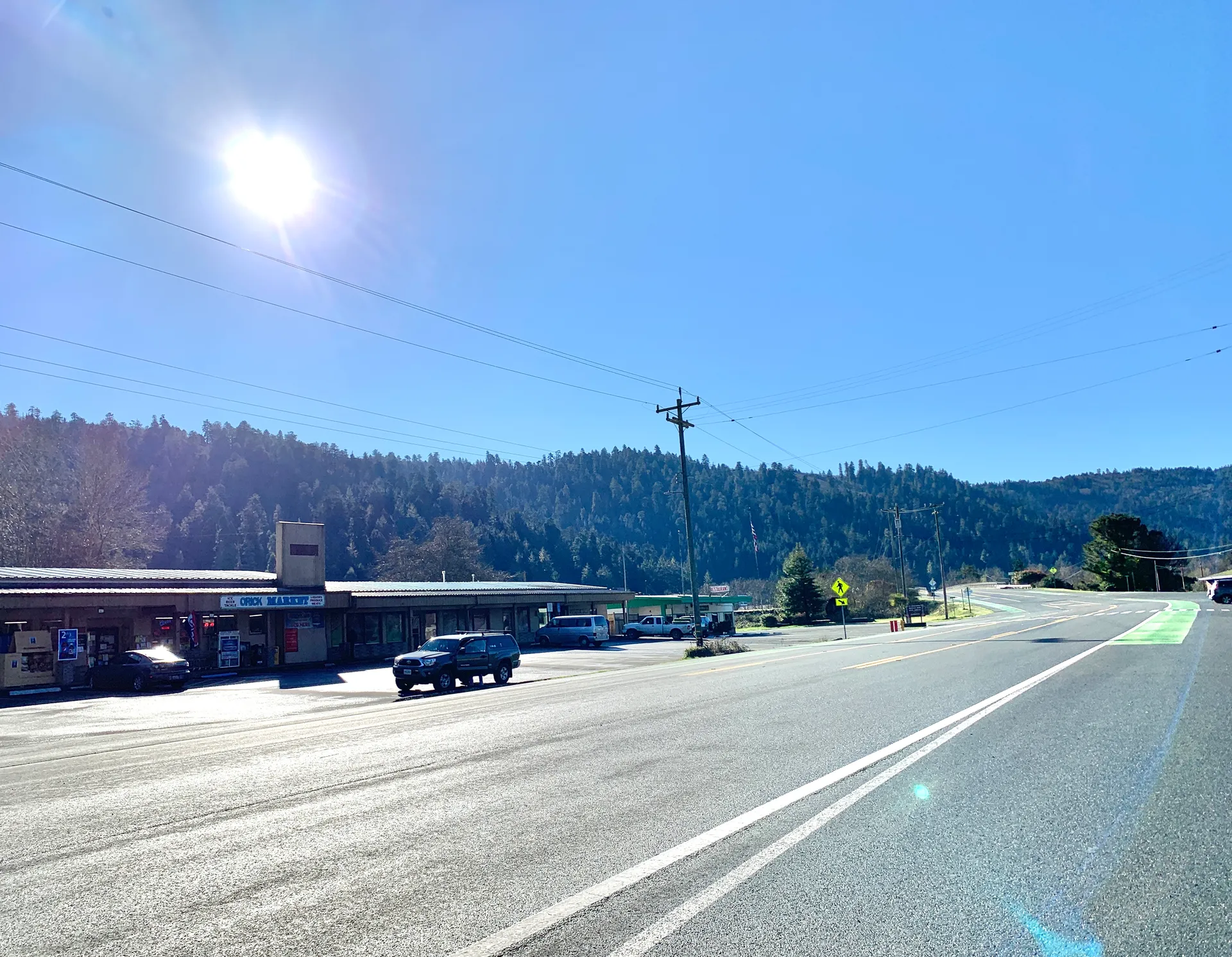 A road and roadside buildings in Orick, California, with forested hills behind them.